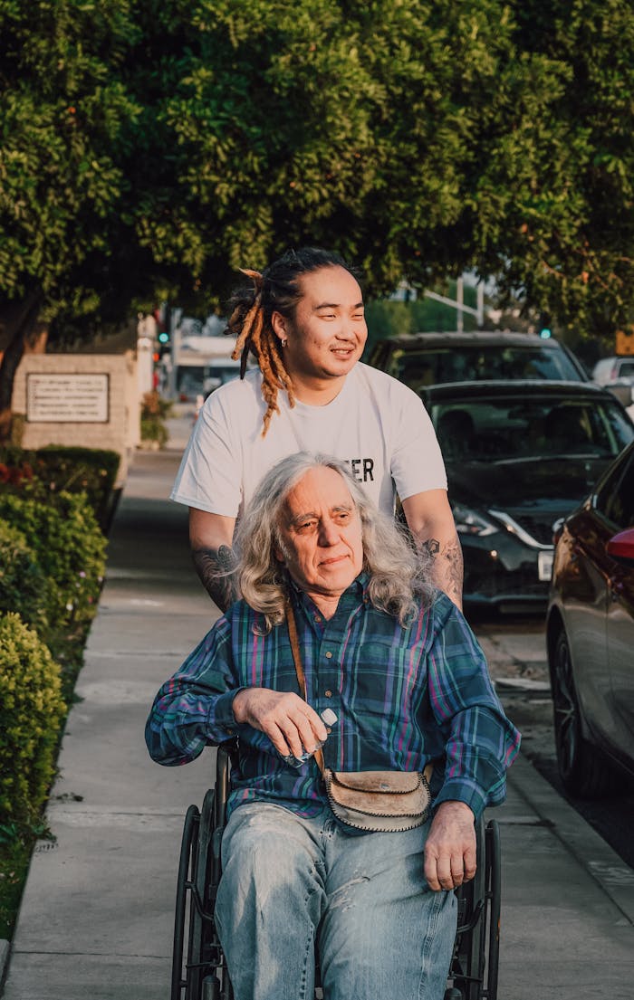 A young man pushes an elderly man in a wheelchair on a sunny sidewalk.