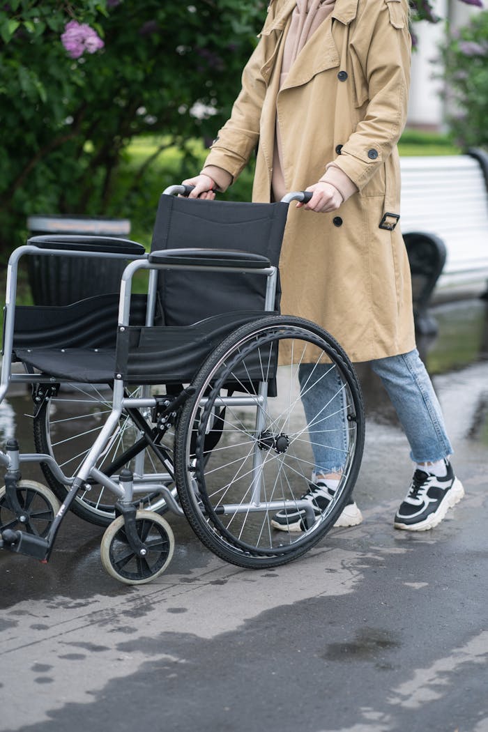 Person wearing a trench coat pushing an empty wheelchair on a wet sidewalk outdoors.